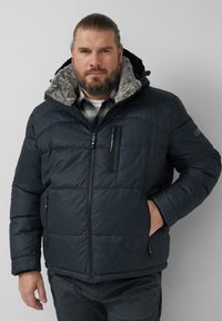 Donkerblauwe pufferjas met een gestructureerde afwerking, ritssluiting en capuchon met nepbontvoering. Voorzien van twee voorzakken en een borstzak met rits.