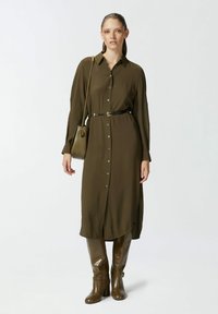 Maslinasta zelena haljina košulja s dugim rukavima i dugmadi sprijeda, s remenom u struku. Uparena s sjajnim smeškim čizmama do koljena. Jednostavna torbica kao dodatak.