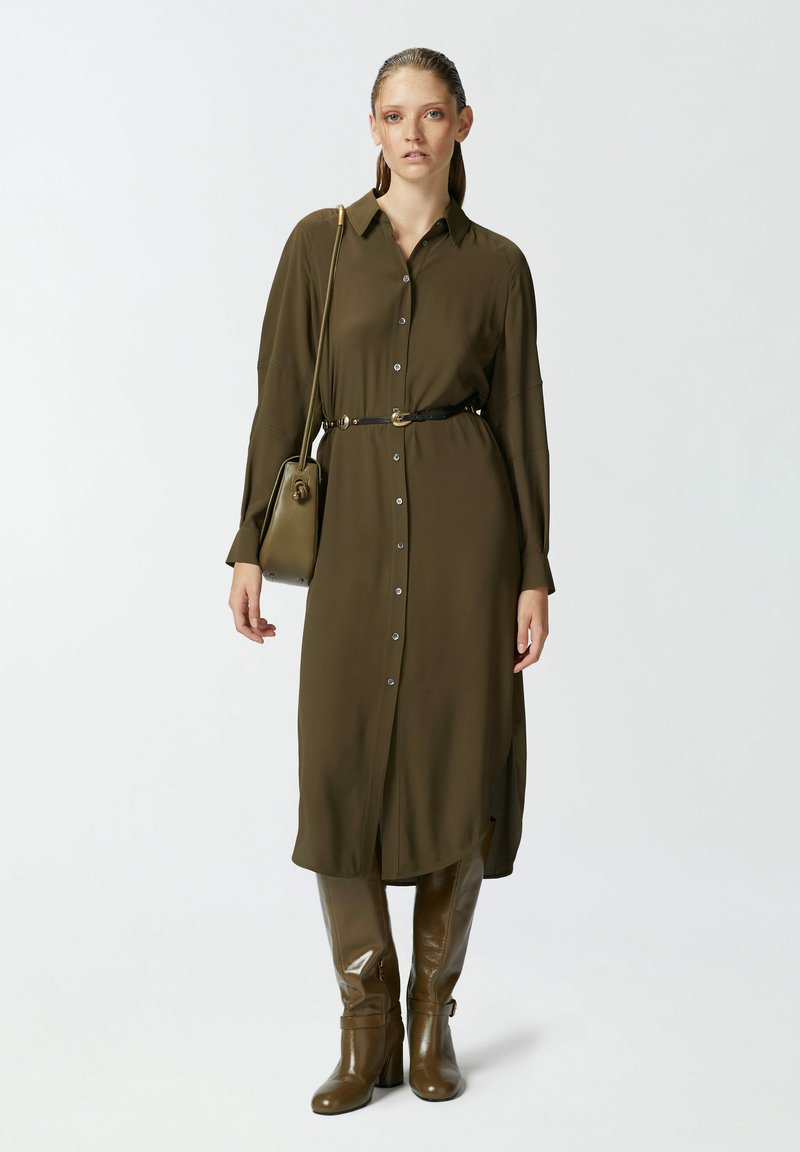 Maslinasta zelena haljina košulja s dugim rukavima i dugmadi sprijeda, s remenom u struku. Uparena s sjajnim smeškim čizmama do koljena. Jednostavna torbica kao dodatak.