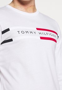 Weißes Langarm-T-Shirt mit einem Grafikdesign, das aus horizontalen Streifen in Schwarz, Rot und Weiß besteht, sowie dem Schriftzug "TOMMY HILFIGER" auf der Brust.