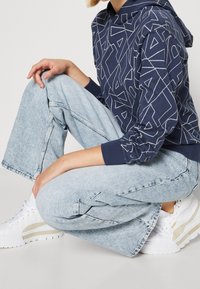 Felpa blu navy con motivo geometrico bianco, jeans a gamba larga azzurri e sneakers bianche con accenti beige. Tessuto in cotone texturizzato.