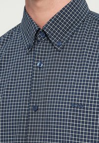 Kék-fehér kockás, gombos ing gallérral, kék gombokkal és hímzett "BOSS" logóval. Közeli kép a szövet textúrájáról.