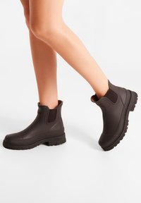 Bottes de cheville en caoutchouc brun avec des semelles texturées, des panneaux latéraux élastiques et une finition lisse. Présentent un talon légèrement surélevé pour le confort.