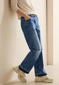 Blaue Jeans aus Denim mit geradem Bein, mit Fronttaschen und Knopfverschluss. Tragen mit hellen Sneakern vor einem neutralen Hintergrund.