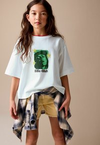 White short-sleeve t-shirt with a green graphic design, featuring a face and stars, paired with yellow shorts and a blue checked flannel tied at the waist.