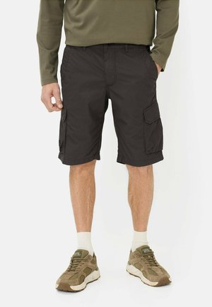 Man draagt donkere cargobroek, olijfgroen shirt met lange mouwen, witte sokken en olijfgroene casual sneakers, staat met één hand in de zak.