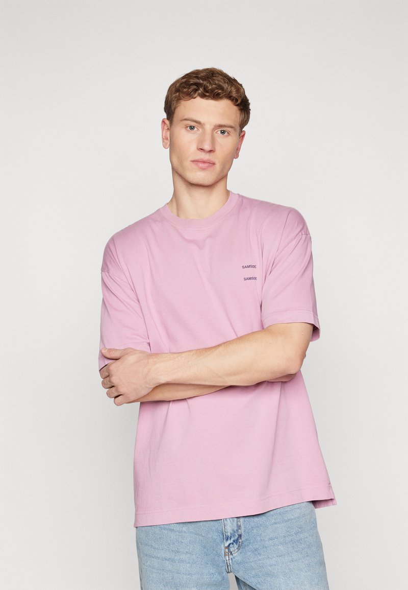 Lyserød bomulds-t-shirt med korte ærmer, rund hals og to små logoer på venstre brystområde; sat sammen med lyseblå denimjeans.