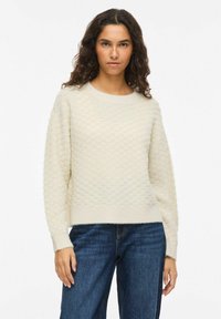 Pull en tricot couleur crème clair avec un motif nid d'abeille texturé, encolure ronde et ourlet côtelé, porté avec un jean bleu foncé.
