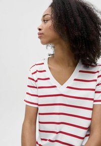 Camiseta de cuello en V a rayas rojas y blancas, confeccionada en tela suave, con mangas cortas y un corte relajado. Los detalles incluyen una costura visible y una textura suave.