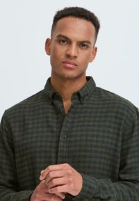 Camisa a cuadros verde y azul marino con cuello abotonado, mangas largas y tela texturizada; dos botones visibles cerca del cuello.