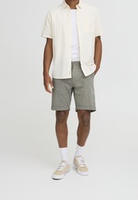 Chemise en lin légère à boutons, t-shirt blanc en dessous, shorts gris à motifs, manches retroussées, baskets crème et chaussettes blanches mi-mollet.