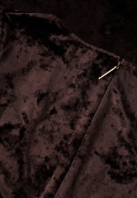 Brunt fløjlstof med et subtilt mønster og en guldzippet accent på siden som lukningsdetalje. Blød tekstur synlig.