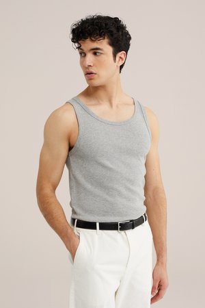 Junger Mann, der ein graues Tanktop und weiße Hose mit schwarzem Gürtel trägt, steht mit einer Hand in der Tasche und schaut zur Seite.