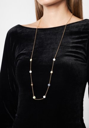 Guld kædetrøje med assorterede runde perler i hvid, grøn, lyseblå og guld. Bæres over en sort fløjlstop, lang længde.