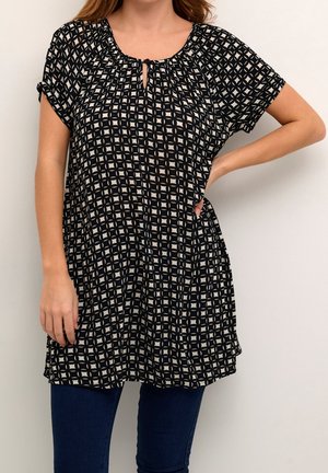 Frau trägt eine schwarze Tunika mit weißem geometrischem Muster und Schlüssellochausschnitt, kombiniert mit dunkelblauen Jeans, steht mit einer Hand in der Hüfte.