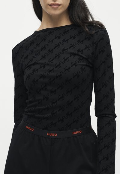 Femme portant un haut noir ajusté à manches longues avec un texte répété "Hugo" et un pantalon noir avec une taille élastique portant le logo "Hugo".