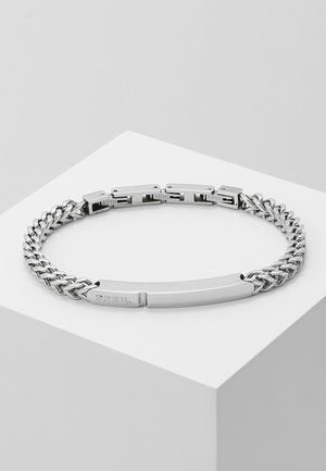 Bracelet chaîne en acier inoxydable argenté avec fermoir rectangulaire et plaque lisse gravée, présenté sur un socle cubique blanc.