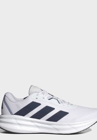 Chaussure de course blanche avec un design à trois bandes noires, semelle rembourrée estampillée Cloudfoam, et lacets blancs sur un fond gris clair uni.