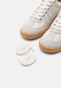 Beige and white sneakers with gum soles, partially visible, next to two neatly coiled white shoelaces on a white background.