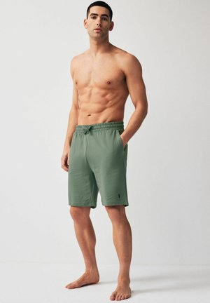 Shorts en coton verts avec une ceinture élastique, des poches latérales et un petit logo en accent. Pas de motifs visibles. Le modèle se tient pieds nus.
