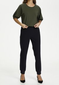 Olivgrön bomullstop med korta, r gathered ärmar kombinerad med mörkblå åtsittande byxor, som har en slät textur och åtsittande design.