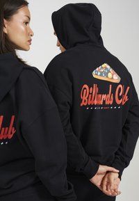 Μαύρες φούτερ διαθέτουν μια μεγάλη γραφική εκτύπωση στην πλάτη, που απεικονίζει ένα τρίγωνο από μπαλάκια μπιλιάρδου και κείμενο που γράφει "Μπιλιάρδος Κλαμπ" σε κόκκινο χρώμα.