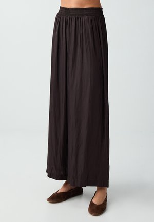 Pantalon noir ample à jambes larges avec taille élastique, porté avec des chaussures plates marron à enfiler sur fond blanc.