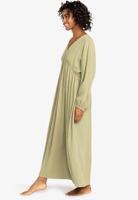 Vrouw met krullend haar die blootsvoets staat, gekleed in een lange, losse, lichtgroene jurk met lange mouwen en een V-hals.