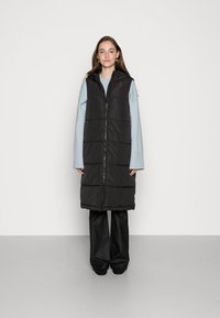 Gilet noir long matelassé avec fermeture éclair, capuche et design matelassé. Porté sur un haut bleu clair et un pantalon large noir.