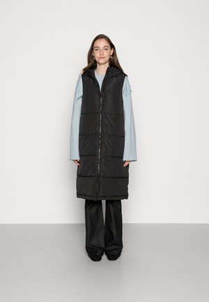 Langer schwarzer gefütterter Weste mit Reißverschluss, Kapuze und gestepptem Design. Getragen über einem hellblauen Oberteil und einer schwarzen weiten Hose.