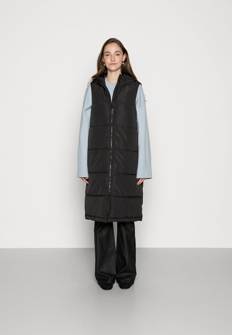 Gilet noir long matelassé avec fermeture éclair, capuche et design matelassé. Porté sur un haut bleu clair et un pantalon large noir.
