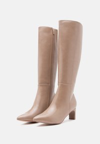 Botas de cuero beige hasta la rodilla con punta afilada y tacón cuadrado. Presentan un zipper lateral y una textura suave con un detalle de costura mínimo.