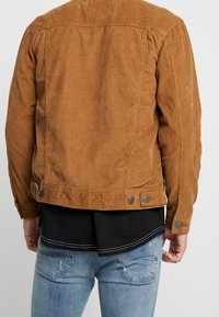 Brązowa kurtka ze sztruksu o teksturowanym wykończeniu, z guzikami w talii i na mankietach, stylizowana z czarną koszulą i jasnoniebieskimi dżinsami.