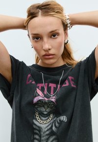 Sort bomulds t-shirt med et grafisk motiv af en kat iført en pink bandana og guld halskæde, med ordet "ATTITUDE" i lilla bogstaver ovenfor.