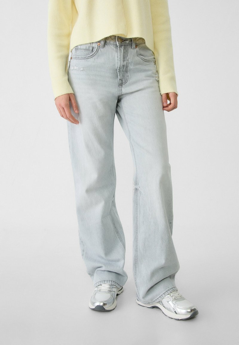 Personne portant un jean large bleu clair, des baskets argentées et blanches, et un haut à manches longues jaune pâle, debout sur un fond uni.