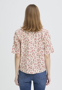 Blouse fleurie en tissu crème ornée de fleurs rouges et bleues, manches courtes et ourlet arrondi. Portée avec un jean bleu, vue de dos.