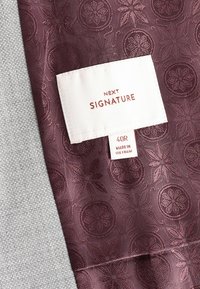 Tissu bourgogne avec motif floral en relief à côté d'un tissu gris texturé ; comprend une étiquette blanche avec "NEXT SIGNATURE" et les détails de taille.