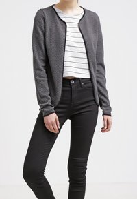 Šedý texturovaný cardigan s kulatým výstřihem a černými detaily, nošený přes bílý pruhovaný top a spárovaný s černými skinny džínami.