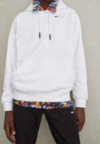White hooded sweatshirt with a colorful, patterned interior collar. Features front kangaroo pocket and drawstrings with black tips. Paired with black pants.