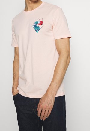 Homme portant un t-shirt à manches courtes rose clair avec un motif vague bleue et soleil rouge sur la poitrine gauche, assorti à un jean foncé.