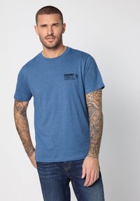 Blå kortärmad bomulls-t-shirt med "TOUGH TRAILS" tryckt i svart på bröstet. Slät textur, rund halsringning och avslappnad passform.
