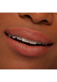 Nærbilde av lepper med en matt, terrakotta leppestiftfarge, jevn tekstur og hvite, jevne tenner, mot en varm hudtone.