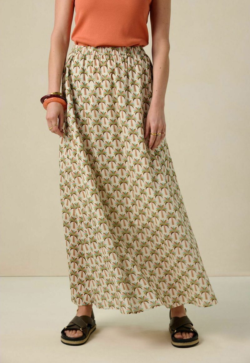 Jupe maxi en tissu beige avec un imprimé de palmier en vert et marron. Taille élastique et silhouette fluide. Associée à des sandales noires.