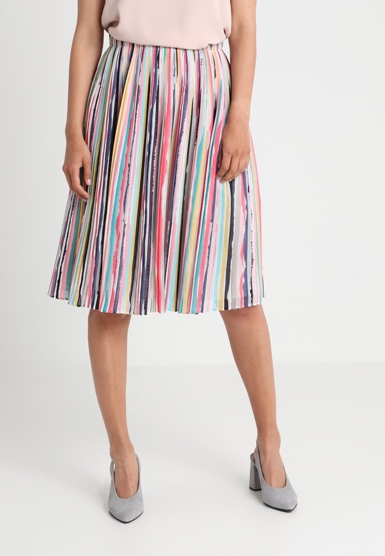 Femme portant une jupe midi plissée avec des rayures multicolores verticales et des chaussures à talons hauts gris clair sur un fond uni.