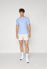 Világoskék atlétikai póló rövid ujjakkal és testhezálló kialakítással, világos bézs rövidnadrággal és fehér tornacipővel. Simább anyag, minimális textúra.