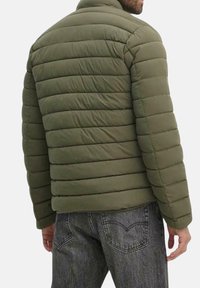 Giacca a vento verde con motivo a righe orizzontali, colletto alto e spalle leggermente arrotondate. Indossata sopra un paio di jeans scuri.