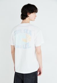 Weißes Baumwoll-T-Shirt mit einem pastellfarbenen Grafikdesign auf der Rückseite, das den Text "SONNE, MEER UND LEE" sowie eine Sonne- und Wellenillustration zeigt.