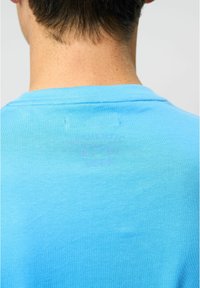 Blå bomulds t-shirt med rund halsudskæring, med et diskret print på ryggen. Stoffet er glat og let rynket.