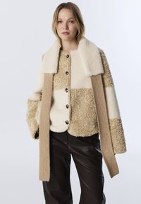 Beige en crème colbert met een textuur van pluche en mouwen van gebreid materiaal met duimgaten. Bevat grote knopen en heeft een boxy pasvorm.