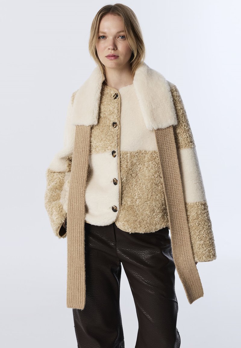 Beige en crème colbert met een textuur van pluche en mouwen van gebreid materiaal met duimgaten. Bevat grote knopen en heeft een boxy pasvorm.